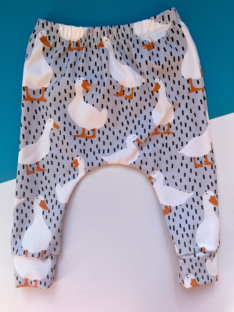 A pair of child harem pants on a blue and white background. Pant fabric print has a grey background with black dashes and a white duck with orange beak and feet.