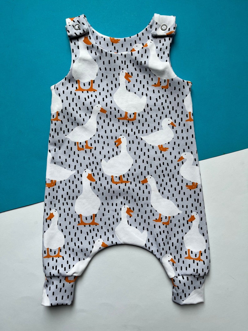 A child's romper on a blue and white background. Romper fabric pattern has a grey background with black dashes and a white duck with orange beak and feet.