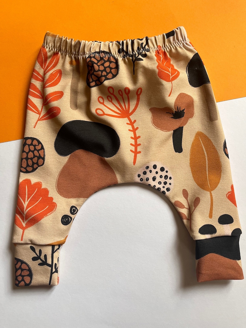Baby trousers on an orange and white background. Trouser fabric has orange and brown mushrooms and leaves all over