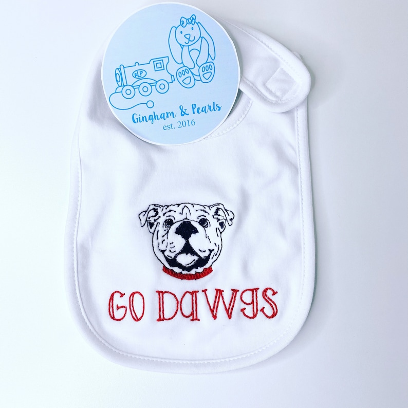 May include: White baby bib with an embroidered bulldog face and the text "GO DAWGS" in red thread. The bib has a snap closure and is made by Gingham & Pearls.