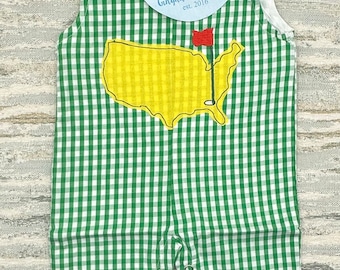 Op de afbeelding: Groen-witte gingham baby-overall met een gele geborduurde omtrek van de Verenigde Staten met een rode golfvlag. De overall heeft een wit label met de tekst "Gingham & Pearls est. 2016".