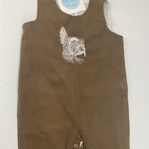 May include: Brown corduroy overalls with a white embroidered chicken design on the front. The overalls have adjustable straps and snap closures at the bottom.