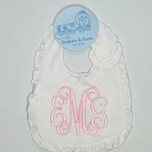 May include: White baby bib with a pink embroidered monogram letter "M" and a ruffled edge. The bib has a tag with a blue illustration of a train and a bunny, and the text "Gingham & Pearls est. 2016".