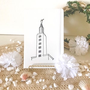 May include: A white card with a black line drawing of a tall building with a statue on top. The card is on a gold stand and surrounded by white flowers and white rocks.