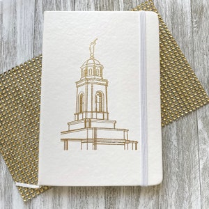 May include: Ivory-colored notebook with a gold-colored illustration of a building on the cover. The notebook has a matching elastic band closure. The background is a light gray wooden surface with a gold-colored, sparkly decorative element.