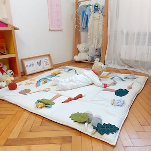 以下が含まれることがあります： クリーム色のプレイマット。木、キツネ、虹など、さまざまなフェルトの形があります。白いロンパースを着た子供がマットの上にいます。子供服の木製ラックとピンクの身長計が背景にあります。