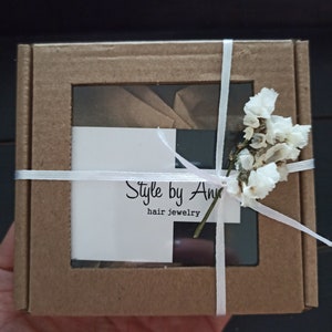May include: A small brown cardboard box with a clear window, tied with white ribbon and decorated with white flowers. The box has a white label with the text "Style by Ann hair jewelry".
