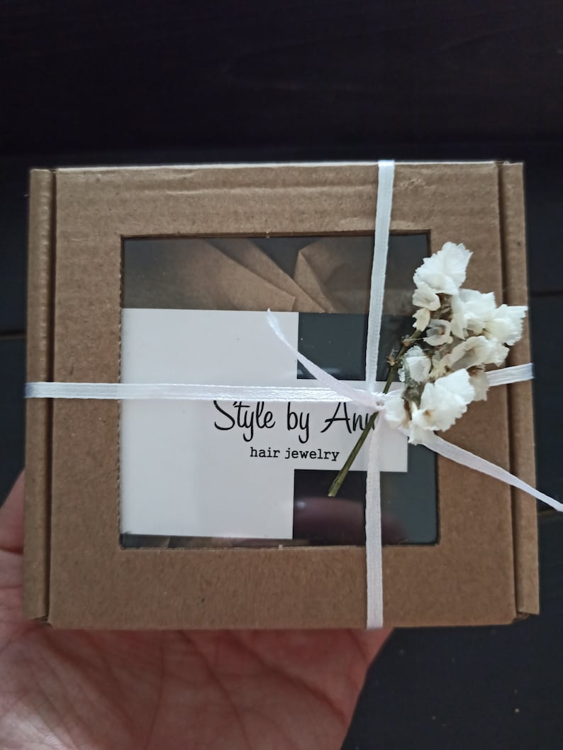May include: A small brown cardboard box with a clear window, tied with white ribbon and decorated with white flowers. The box has a white label with the text "Style by Ann hair jewelry".