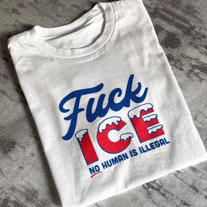 May include: White t-shirt with the text "Fuck ICE" in blue script and red block letters. The text "NO HUMAN IS ILLEGAL" is printed below in a smaller font. The shirt is folded on a gray surface.