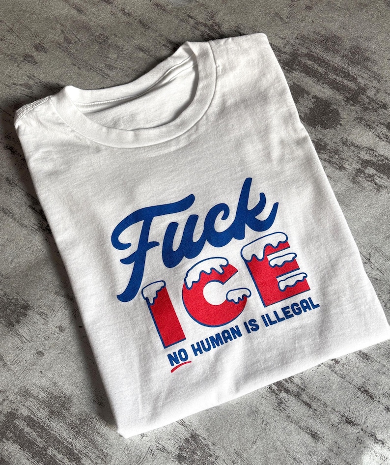 May include: White t-shirt with the text "Fuck ICE" in blue script and red block letters. The text "NO HUMAN IS ILLEGAL" is printed below in a smaller font. The shirt is folded on a gray surface.