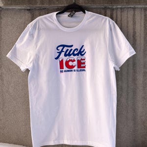 May include: White t-shirt with the text "Fuck ICE" in blue script and red block letters, with the words "NO HUMAN IS ILLEGAL" below. The shirt is hanging on a black hanger against a gray wall.
