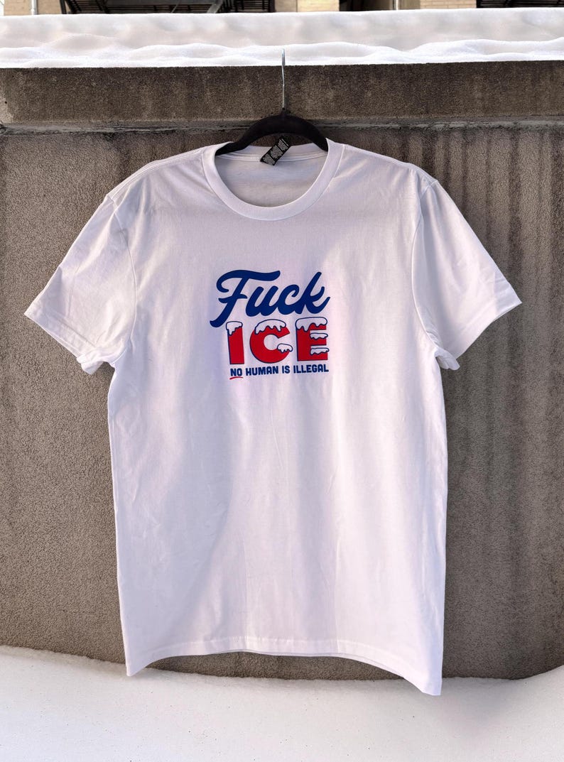 May include: White t-shirt with the text "Fuck ICE" in blue script and red block letters, with the words "NO HUMAN IS ILLEGAL" below. The shirt is hanging on a black hanger against a gray wall.