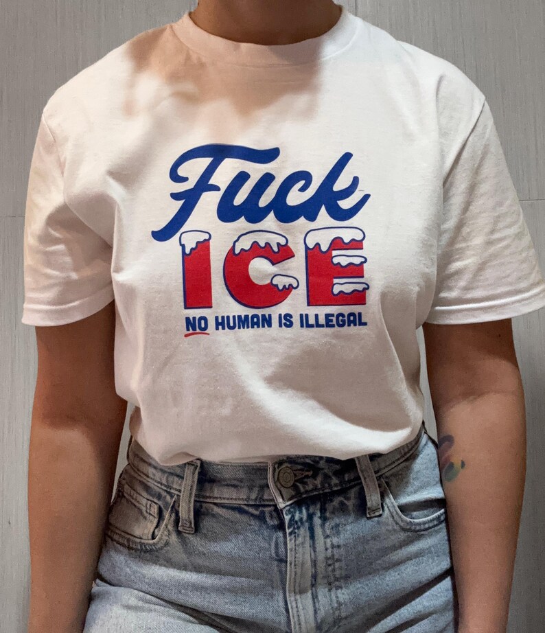 May include: White t-shirt with the words "Fuck ICE" in blue script and red block letters. The text "NO HUMAN IS ILLEGAL" is printed below in red. The shirt is made of a soft material.