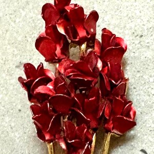 May include: A brooch featuring a bouquet of deep red enameled roses with layered petals. The stems and leaves are crafted from a textured gold-tone metal, forming a long, slender base that supports the cluster of vibrant red flower heads at the top.