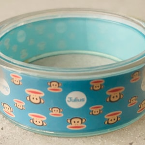May include: A clear plastic bangle bracelet with a blue background and a repeating pattern of brown monkeys with red cheeks. The text "Julius" is printed in white on the bracelet.