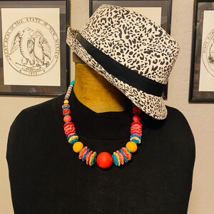 May include: A black turtleneck shirt with a colorful beaded necklace and a black and white leopard print fedora hat. The necklace features red, yellow, and blue beads. The hat has a black band.