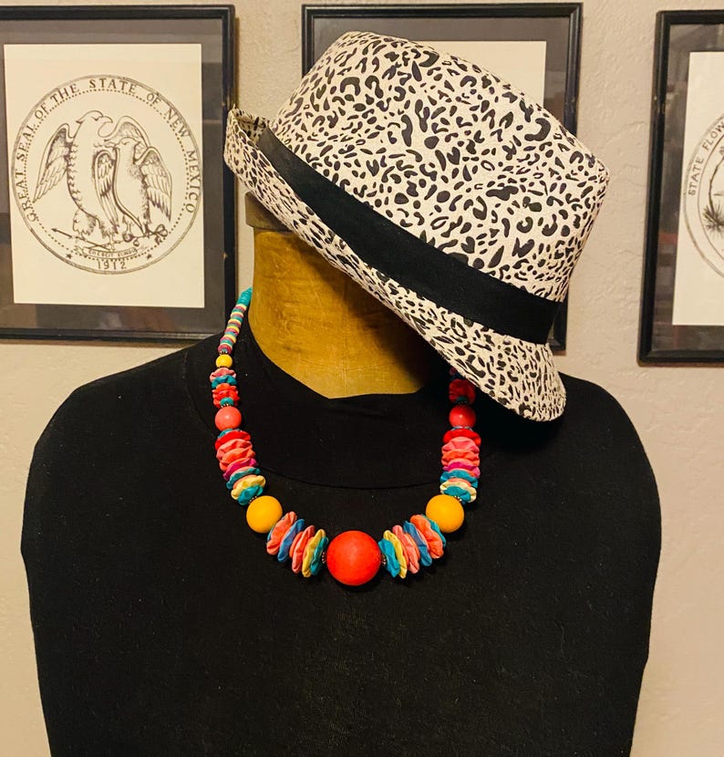 May include: A black turtleneck shirt with a colorful beaded necklace and a black and white leopard print fedora hat. The necklace features red, yellow, and blue beads. The hat has a black band.