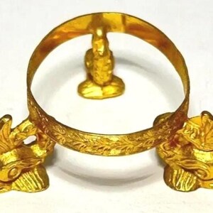 May include: A gold-colored decorative stand with a circular design, featuring two ornate animal figures at the base and a small figure at the top. The stand has a detailed, engraved pattern around the circular frame.