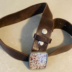 May include: A brown leather belt with a silver buckle featuring a floral design. The buckle is square shaped and has a detailed floral pattern.