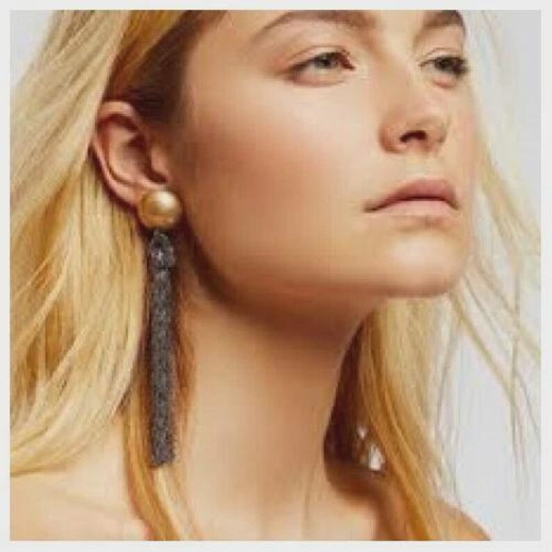 K&ouml;nnte beinhalten: Ein goldfarbener Ohrstecker mit einer langen, dunkelgrauen Quaste. Der Ohrring wird am linken Ohr getragen. Die Frau hat blondes Haar und helle Haut.