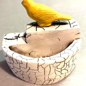 May include: A small, decorative planter featuring a bright yellow bird figurine perched on top of a white, semi-circular base. The base has a distinct black crackle pattern across its surface, giving it a textured, weathered appearance.