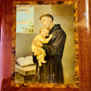 May include: A framed image of a religious painting depicting a man in a brown robe holding a young child. The man is wearing a rosary and the child is wearing a yellow dress. The painting is set against a background of a window with a view of a cloudy sky.