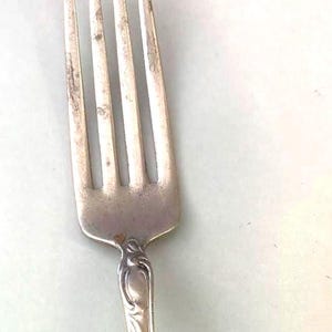 May include: A silver fork with four tines, showing signs of wear. The handle features an ornate, engraved design. The fork is a classic piece of silverware, suitable for dining.