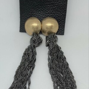 K&ouml;nnte beinhalten: Ein Paar goldfarbene Ohrstecker mit langen, dunkelgrauen Kettenquasten. Die Ohrringe sind an einer schwarzen Kunstlederkarte mit einem einzelnen Loch oben befestigt. Der Hintergrund ist wei&szlig;.