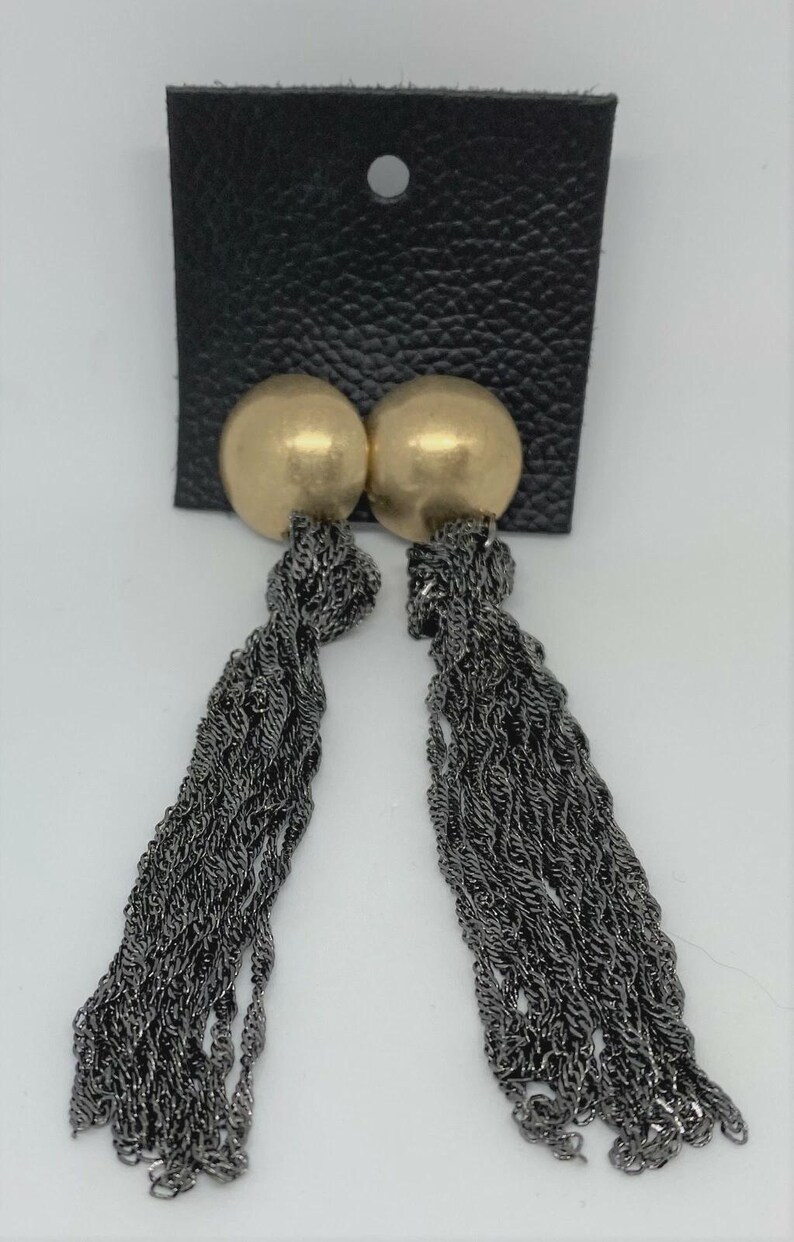 K&ouml;nnte beinhalten: Ein Paar goldfarbene Ohrstecker mit langen, dunkelgrauen Kettenquasten. Die Ohrringe sind an einer schwarzen Kunstlederkarte mit einem einzelnen Loch oben befestigt. Der Hintergrund ist wei&szlig;.