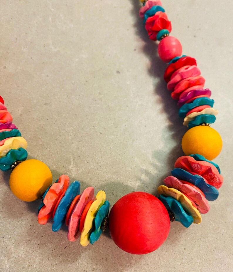 May include: A colorful necklace featuring a variety of beads. The necklace includes large, round, yellow and red beads, along with smaller, flower-shaped beads in shades of pink, blue, and yellow. The necklace is strung on a cord.
