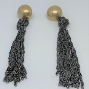 K&ouml;nnte beinhalten: Ein Paar Ohrh&auml;nger. Jeder Ohrring hat einen goldfarbenen, runden Stecker und eine lange, kaskadierende Quaste aus dunkelgrauen Metallketten. Die Ketten sind eng miteinander verwoben und erzeugen einen strukturierten Effekt.