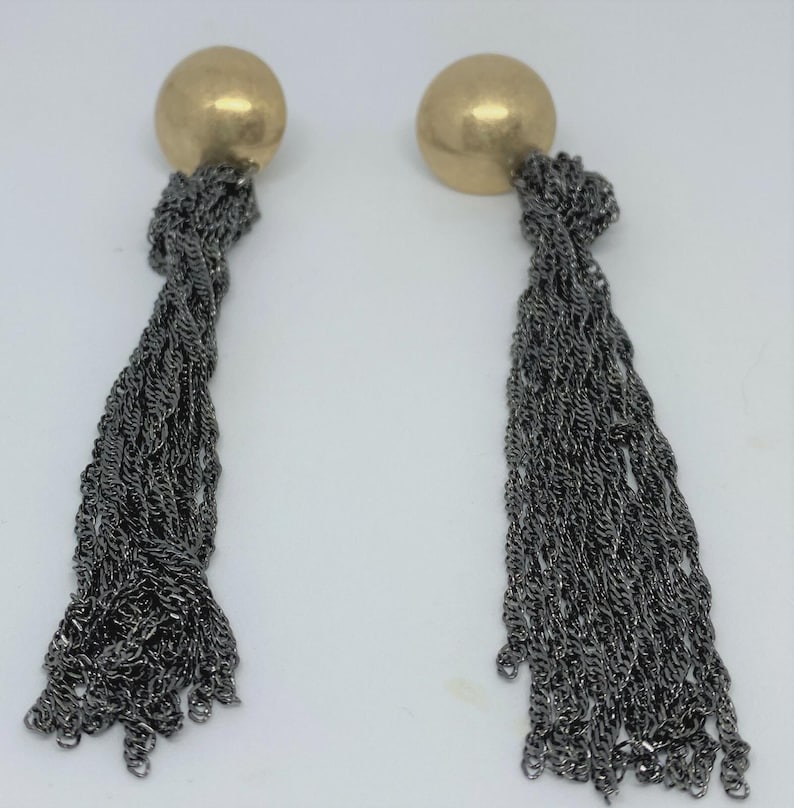 K&ouml;nnte beinhalten: Ein Paar Ohrh&auml;nger. Jeder Ohrring hat einen goldfarbenen, runden Stecker und eine lange, kaskadierende Quaste aus dunkelgrauen Metallketten. Die Ketten sind eng miteinander verwoben und erzeugen einen strukturierten Effekt.