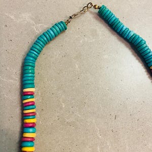 May include: A turquoise beaded necklace with a gold-colored clasp. The necklace features a mix of turquoise, yellow, pink, and white beads. The beads are flat and round, creating a colorful and eye-catching design.