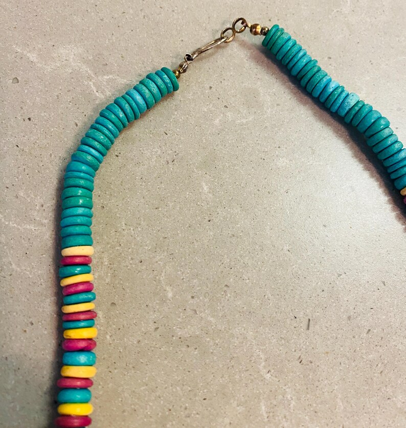 May include: A turquoise beaded necklace with a gold-colored clasp. The necklace features a mix of turquoise, yellow, pink, and white beads. The beads are flat and round, creating a colorful and eye-catching design.