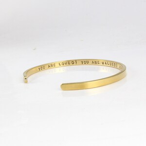 K&ouml;nnte beinhalten: Goldenes Armreif-Armband mit der Inschrift "You are loved! You are valued!"