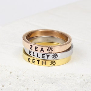 May include: Three stacked rings with engraved names "Zea", "Elley", and "Beth" and a paw print symbol on each ring. The rings are gold, silver, and rose gold.