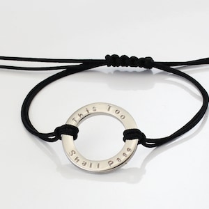 Può includere: Un braccialetto in corda nera con un ciondolo a forma di anello argentato che recita "This Too Shall Pass".
