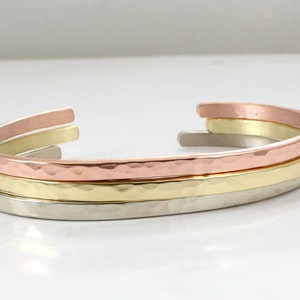 May include: Three stacked bracelets, one silver, one gold, and one copper, with a hammered texture.