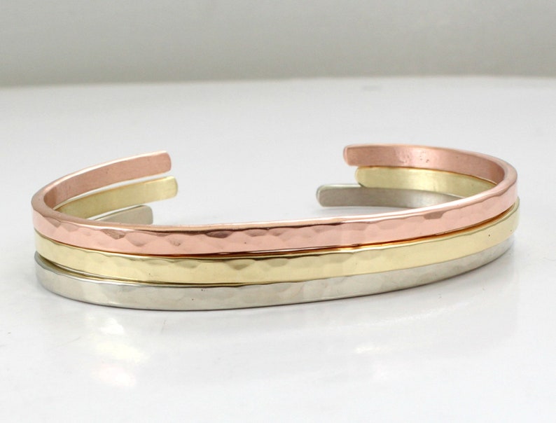 May include: Three stacked bracelets, one silver, one gold, and one copper, with a hammered texture.