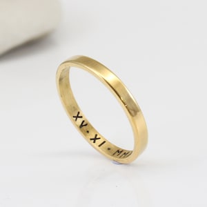 Könnte beinhalten: Ein goldener Bandring mit schlichtem Design. Die Innenseite des Bandes ist mit den römischen Ziffern "XV.XI.MMI" graviert.