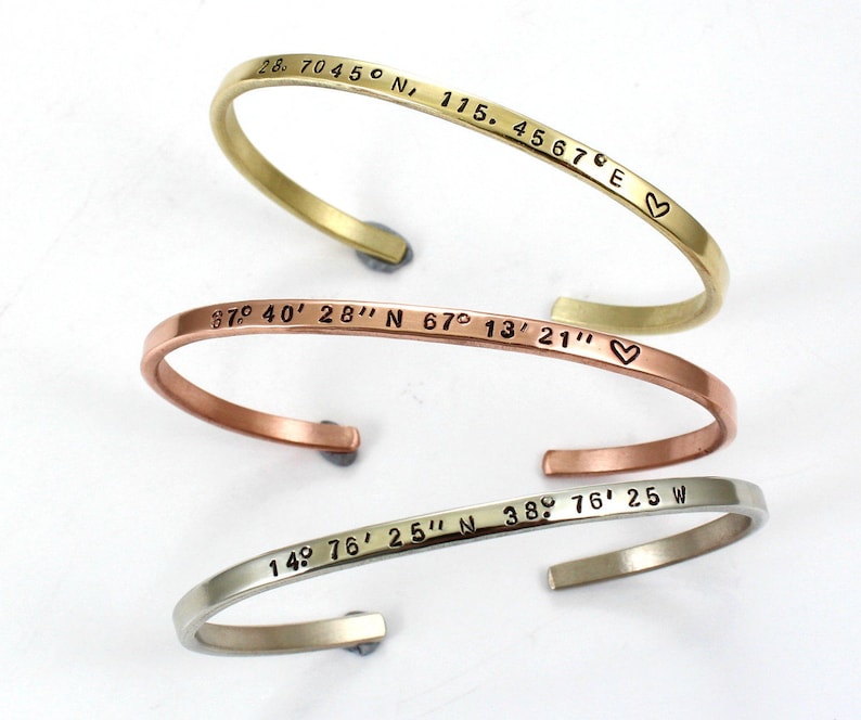 May include: Three gold, copper, and silver bracelets with engraved coordinates and a heart symbol. The coordinates are 28 7045&deg; N, 115.4567E, 87&deg; 40' 28" N 67&deg;13&prime;21&Prime;, and 14&deg; 76'25" N 38 76' 25 W.
