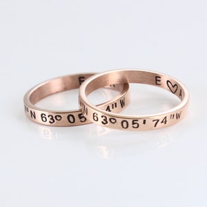 May include: Two rose gold rings with engraved coordinates and a heart symbol. The rings are stacked on top of each other. The coordinates are 41° 05' 74" W and 63° 05' 63° N.