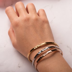 May include: Three hammered metal bangles in gold, silver, and rose gold. The bangles are stacked on a person's wrist.