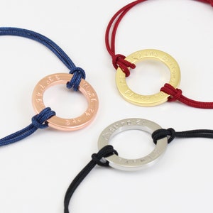 May include: Three bracelets with different colored cords and metal rings. The rings have engraved text. The top bracelet has a red cord and a gold ring with the text "She Believes in Me". The middle bracelet has a blue cord and a rose gold ring with numbers engraved on it. The bottom bracelet has a black cord and a silver ring with the text "Love You To The Moon & Back".