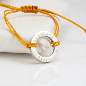 Op de afbeelding: Een geel touw armband met een zilveren ring bedel met de tekst "Be Badass Every Day".