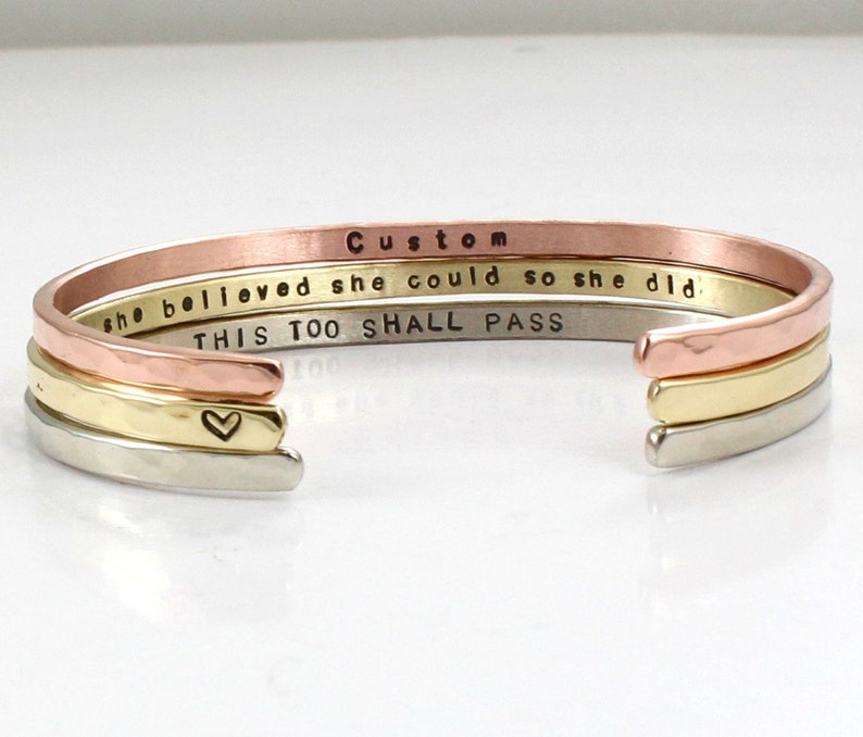 May include: A set of three gold, silver, and copper bracelets with the words "she believed she could so she did" and "this too shall pass" engraved on them.