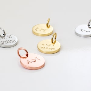 Puede incluir: Cuatro dijes redondos de metal con diferentes grabados. Los dijes son de oro, plata y oro rosa. Los grabados incluyen "A♥T always", "KPE", "XV.VI MMXVIII" y un corazón con "M".