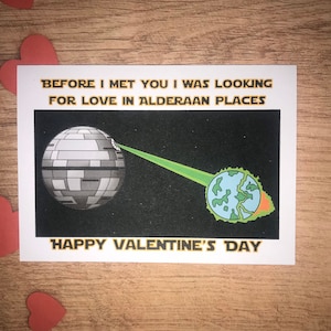 Puede incluir: Tarjeta de San Valentín con una Estrella de la Muerte destruyendo un planeta. La tarjeta dice "BEFORE I MET YOU I WAS LOOKING FOR LOVE IN ALDERAAN PLACES" y "HAPPY VALENTINE'S DAY" en letras doradas. Recortes de corazones rojos.
