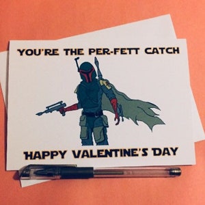 Puede incluir: Tarjeta de San Valentín con una ilustración de dibujos animados de un personaje con casco y capa, sosteniendo un bláster. La tarjeta dice "YOU'RE THE PER-FETT CATCH" y "HAPPY VALENTINE'S DAY". Un bolígrafo descansa debajo de la tarjeta.