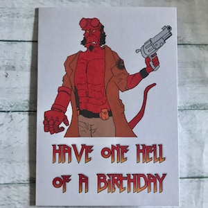 May include: A birthday card with a cartoon illustration of a red demon-like figure holding a gun. The figure wears a brown coat and trousers. The card's text reads "Have One Hell of a Birthday" in orange.